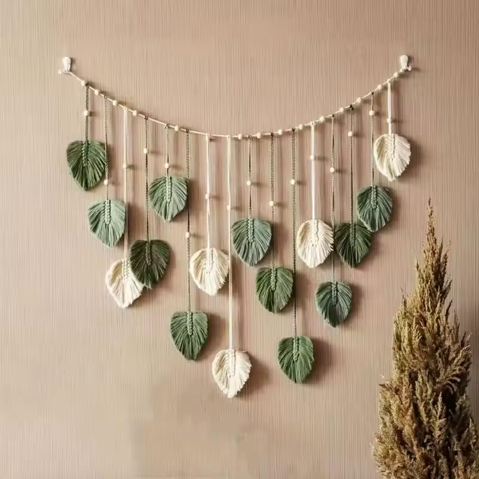 A Plantish Leaf Macrame with green and white leaves and a white string hanging on a textured light brown wall. 