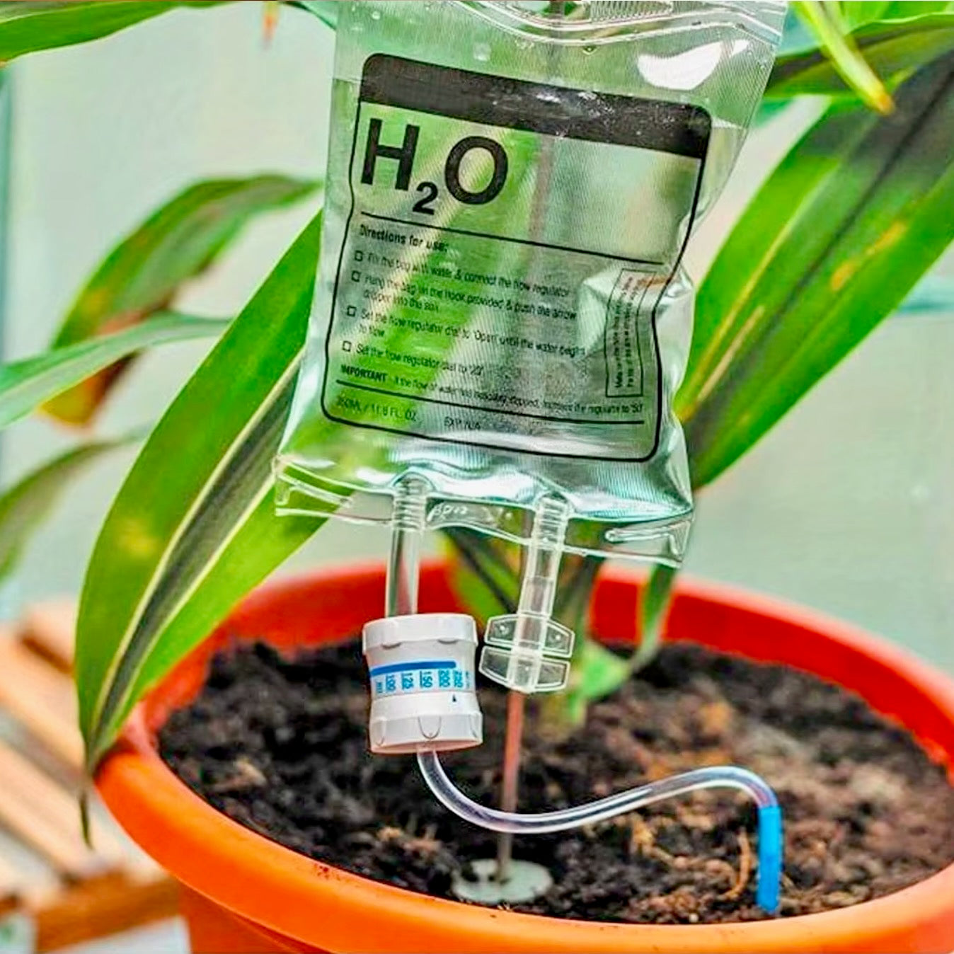 The Plantish IV Self Watering Plant Life Support Bag, shown hanging on a metal hook and placed inside a plastic plant pot full of soil and a green plant.