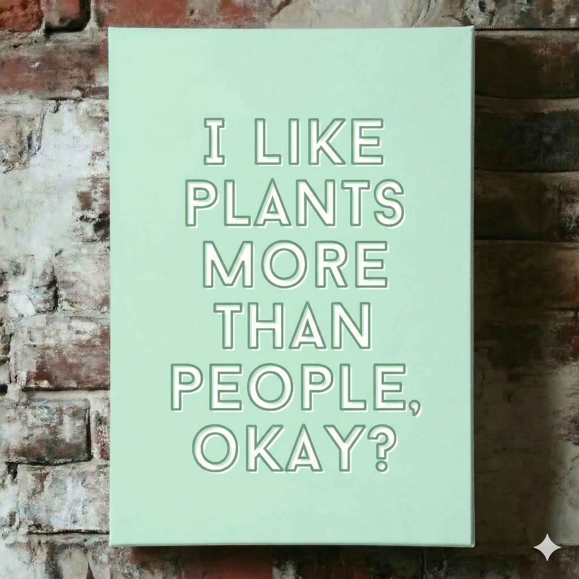 A Plantish canvas of the phrase "I like plants more than people, okay?" in a white font with green background, shown hanging on a red brick wall.