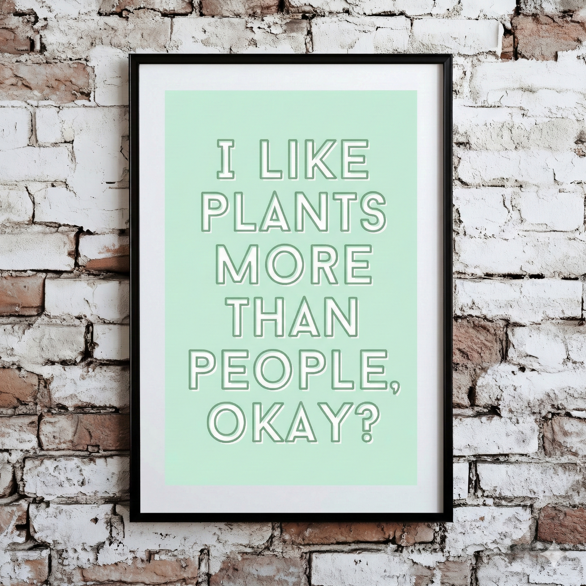 A Plantish print of the phrase "I like plants more than people" in a white font with green background, shown hanging on a brick wall.