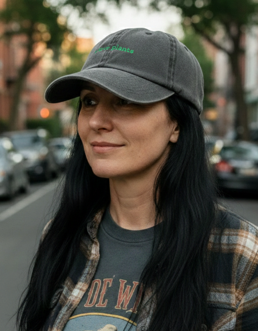 A woman wearing a Plantish I Love Plants Hat in Vintage Charcoal with Green embroidery. She's wearing a check shirt, black band tshirt, and is standing in the street.