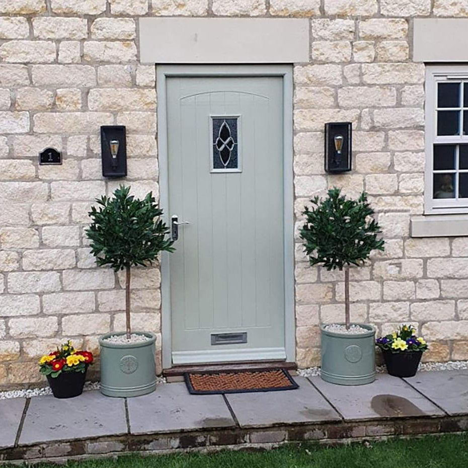 The Plantish Artificial Topiary Laurel Ball Trees, shown in ceramic pots either side of a green cottage door. The green leaves and realistic trunks are displayed. 
