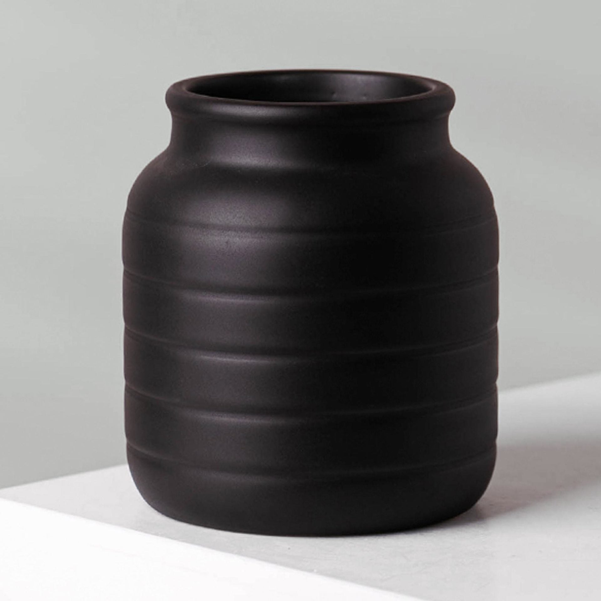 A back close up view of a small ceramic black vase with horizontal groove design. The neck of the vase is slimmer than the bottom.
