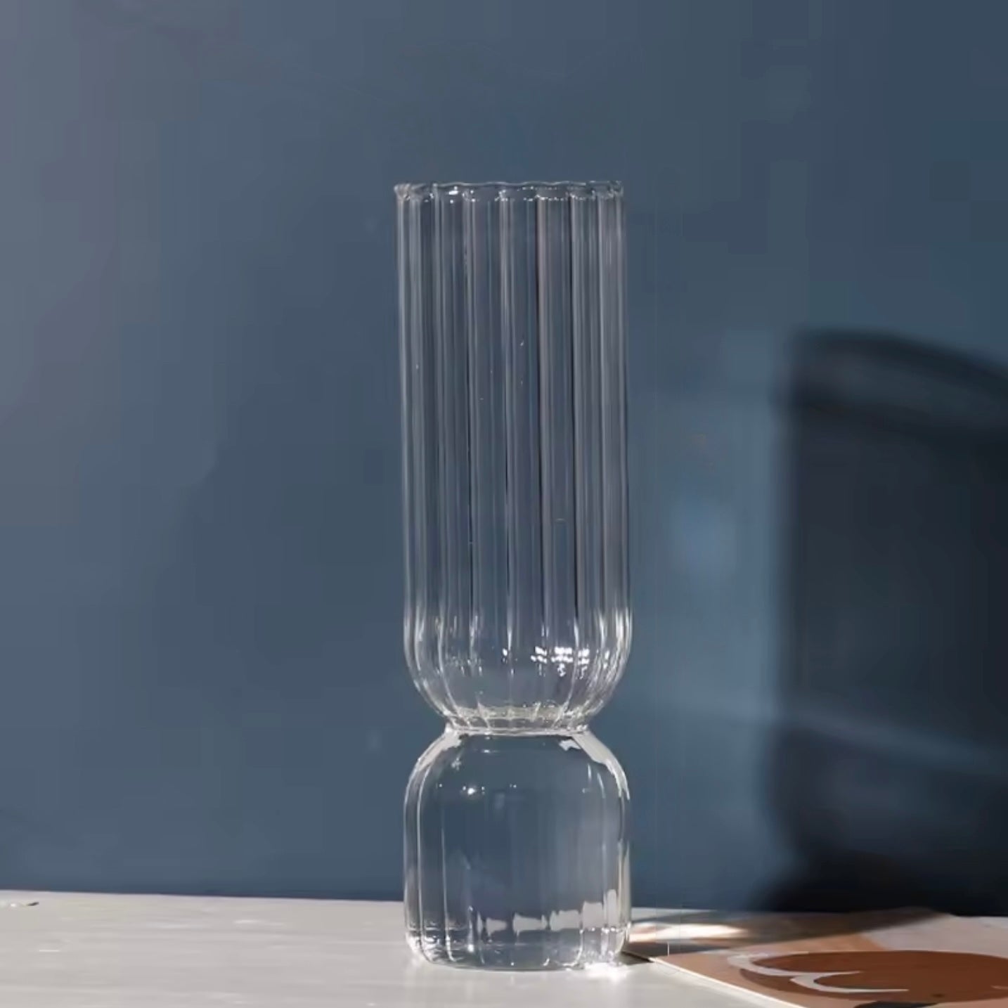 A close up of the Plantish Little Nordic Vase, clear colour option. The vase is on a grey table and there is a mid blue wall in the background.