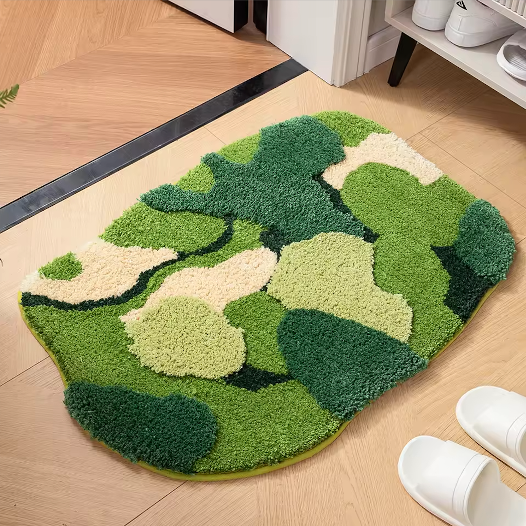 An image of the Plantish Lush Moss Rug, top of the rug and placed on a wooden floor. There is a pair of slippers next to the rug and a shoe rack to the side.
