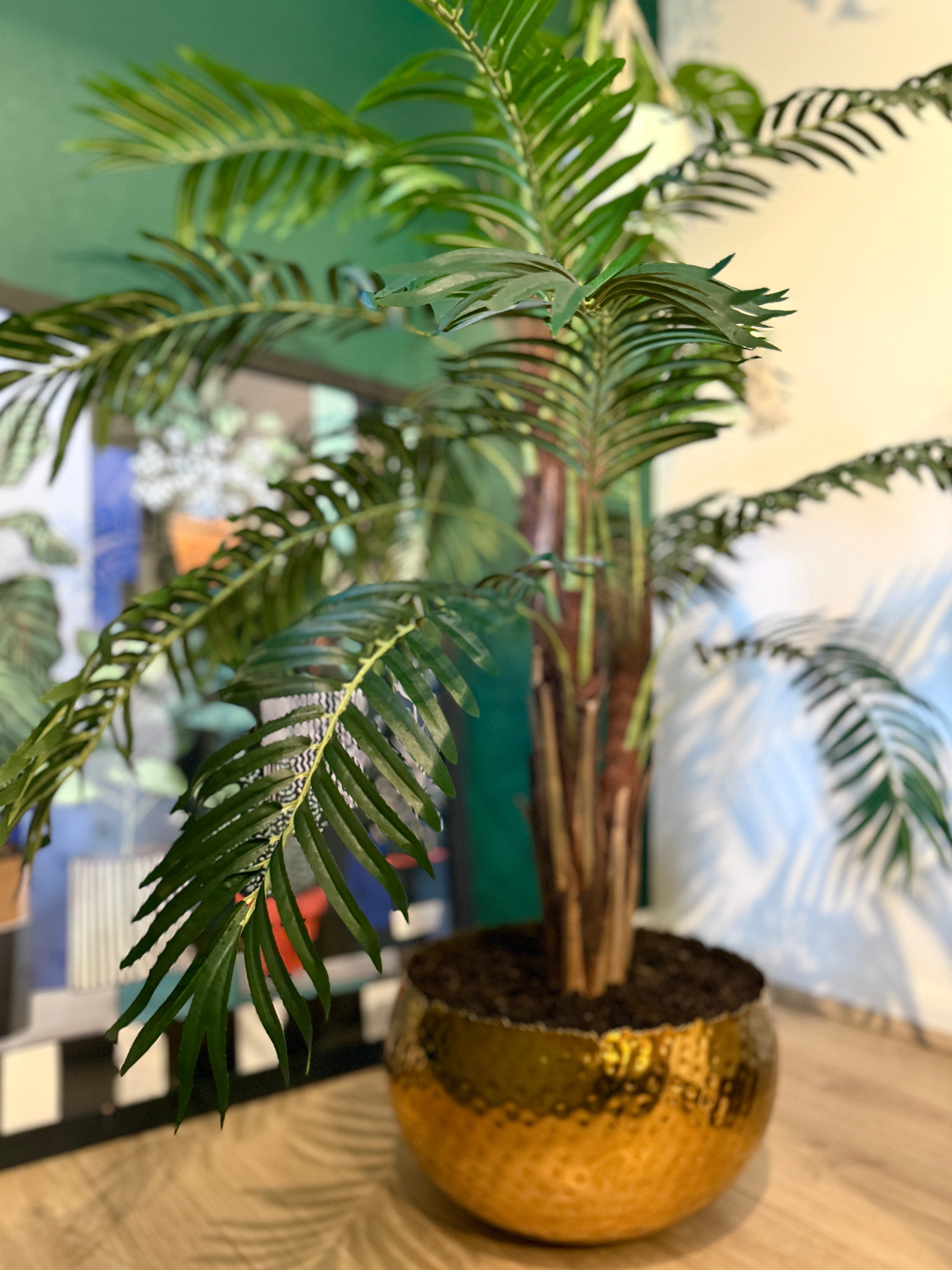 Back view of a Plantish artificial majesty palm, highlighting the layered green leaves, natural bark-covered stem, and hammered gold metal planter filled with real soil.