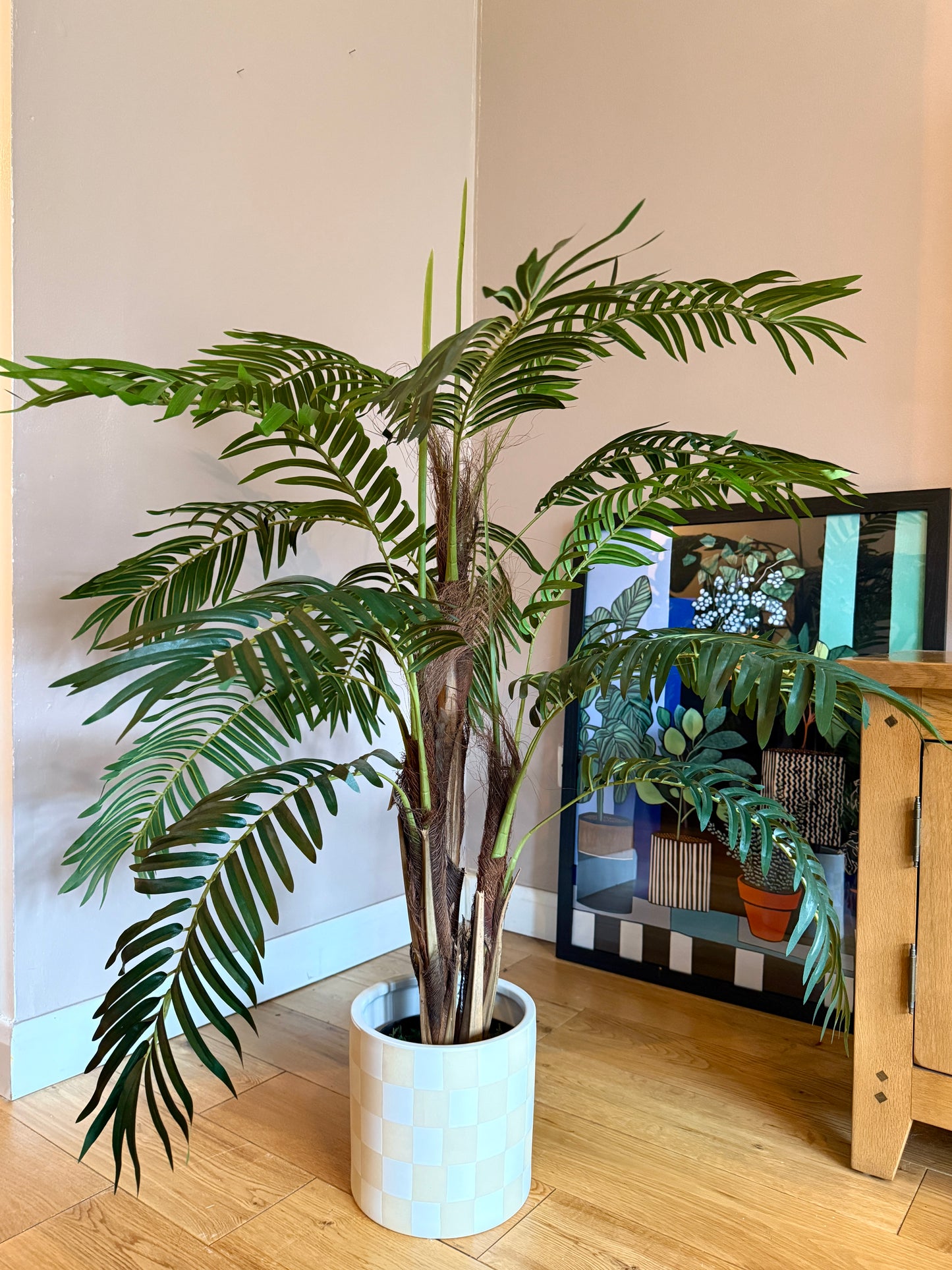 Front view of a Plantish artificial majesty palm with detailed leaves in various shades of green and a natural bark-covered stem, potted in a chequered ceramic pot. 