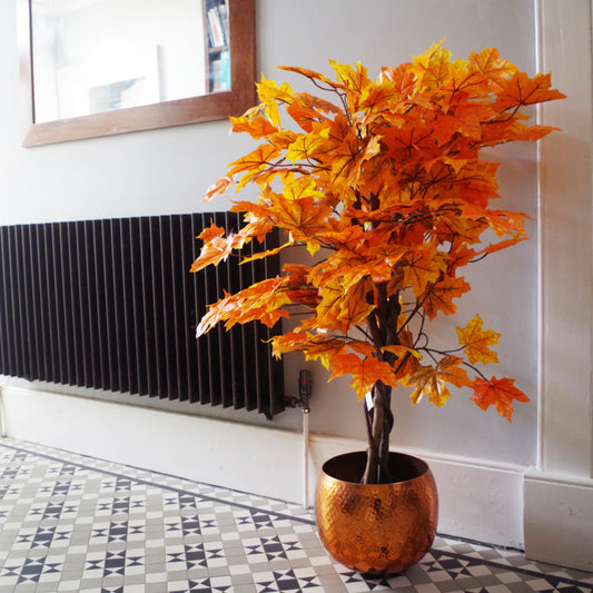 The Plantish artificial maple acer tree shown in a sunlit hallway, highlighting the orange leaves, and inside a copper pot. The floor is a tiled mosaic pattern. 