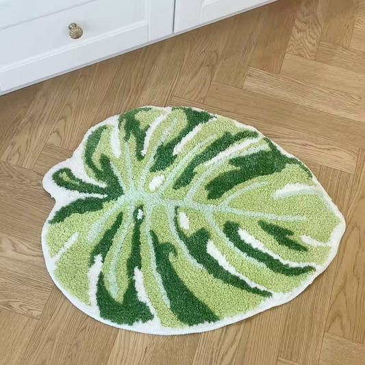 The Plantish Monstera Leaf Tufted Rug positioned on a wooden herringbone floor with a white dresser partially visible behind. The rug is two tones of green and white. 