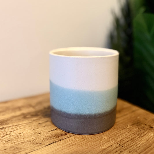 A small ceramic plant pot with an ombre design horizontally, in white, blue, and grey from top to bottom, on a wooden table. 