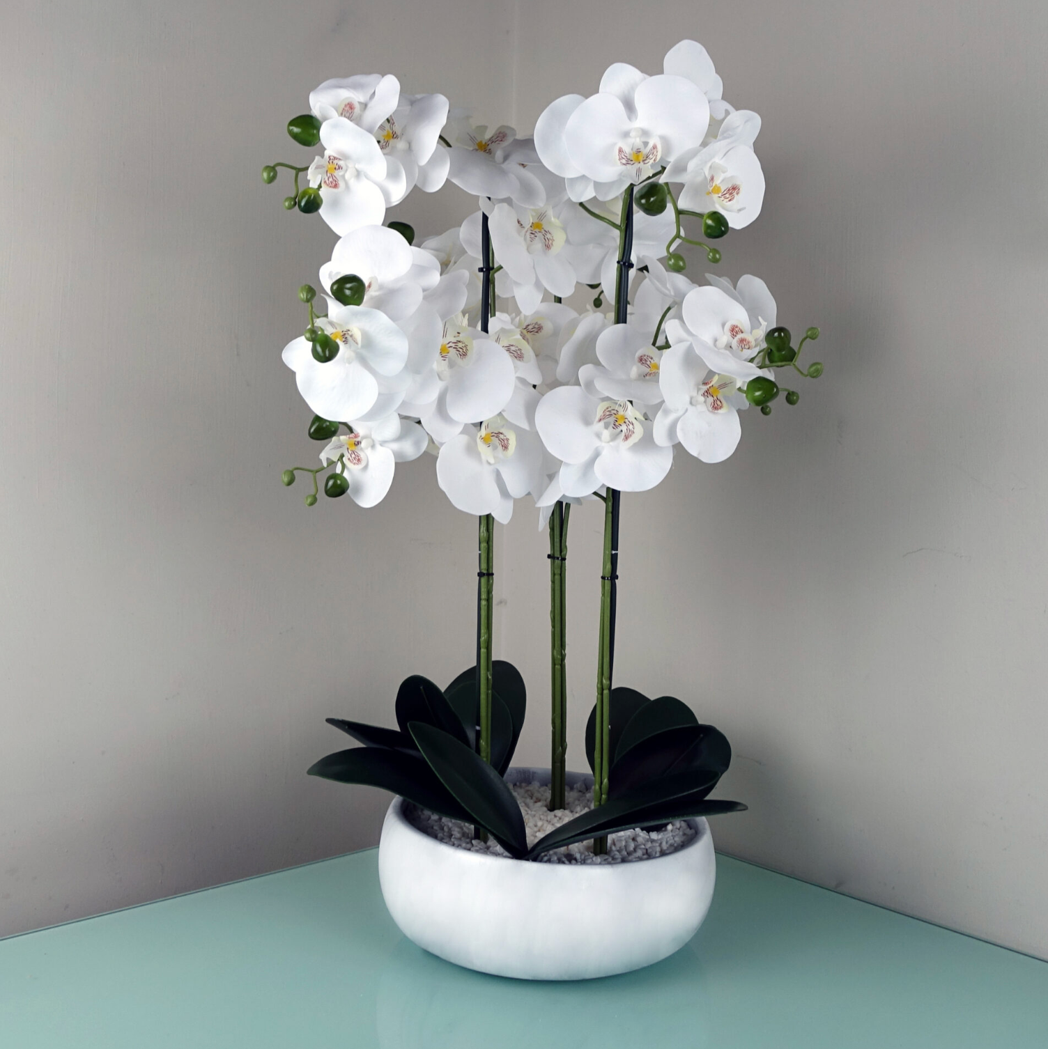An image of the Plantish Artificial Orchid with white flowers, green buds and leaves,  in a white marble effect pot with decorative stones, shown on a table. 