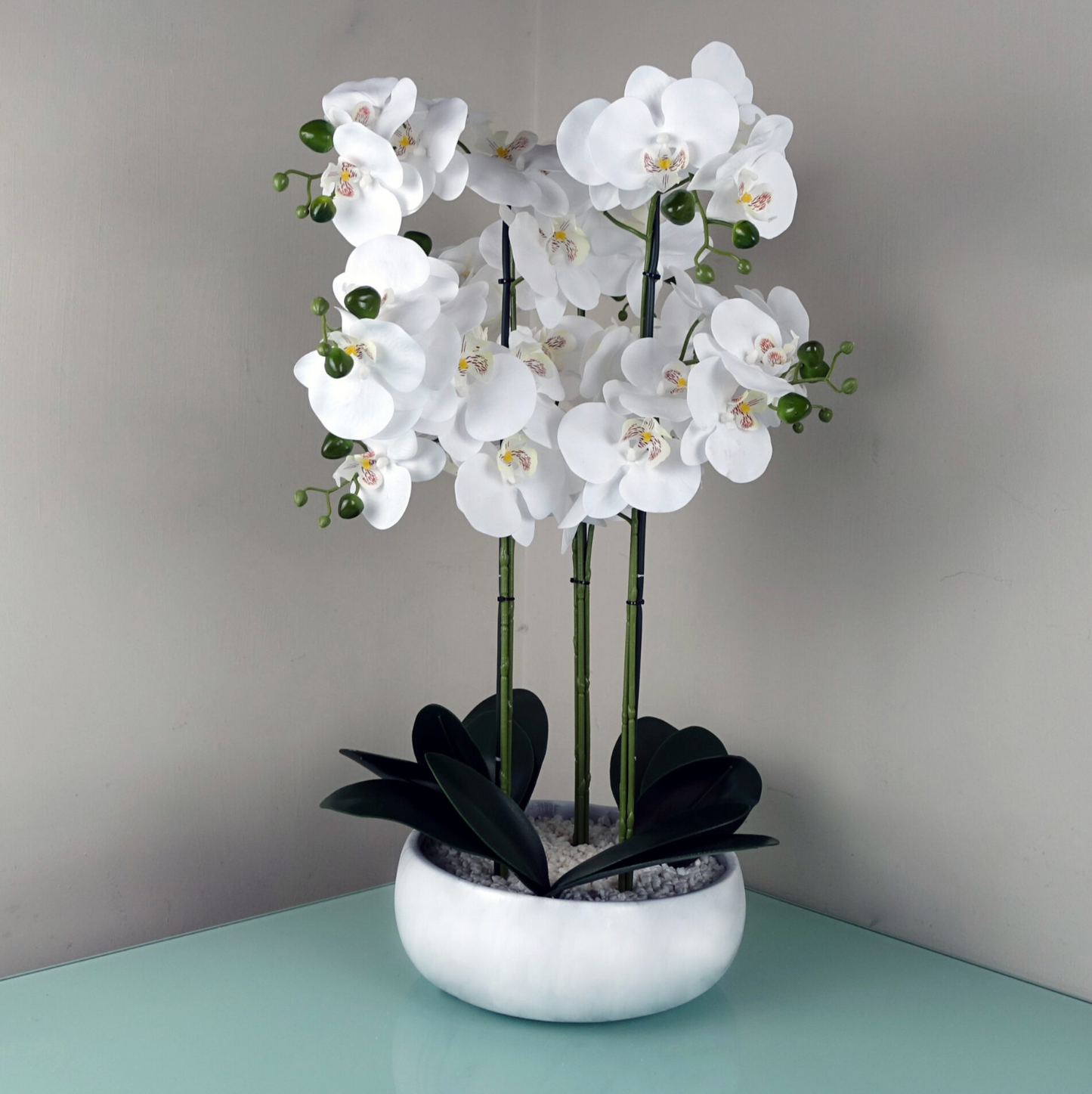 An image of the Plantish Artificial Orchid with white flowers, green buds and leaves,  in a white marble effect pot with decorative stones, shown on a table. 