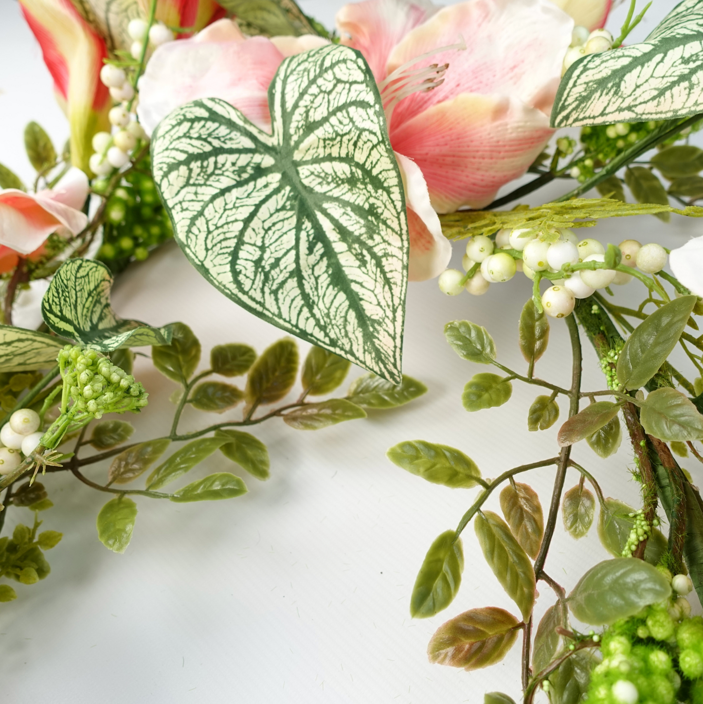 A close up of the Plantish Pink Lily Garland, showcasing variegated alocasia leaves, as well as smaller green leaves and white berries. 