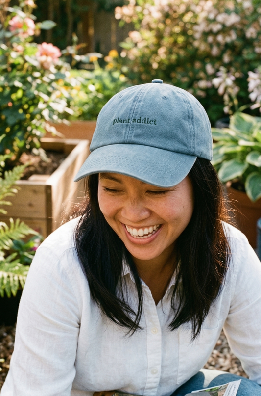 A woman with brown hair wearing a Plantish Plant Addict Hat in Vintage Denim with Green embroidery. She is wearing a white shirt and is crouched in a garden.