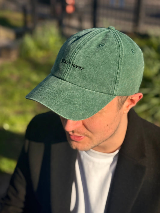 A man wearing a Plantish Plant Lover Hat in Vintage Bottle Green with Black embroidery. He's wearing a black coat, white shirt, and sitting on a park bench.