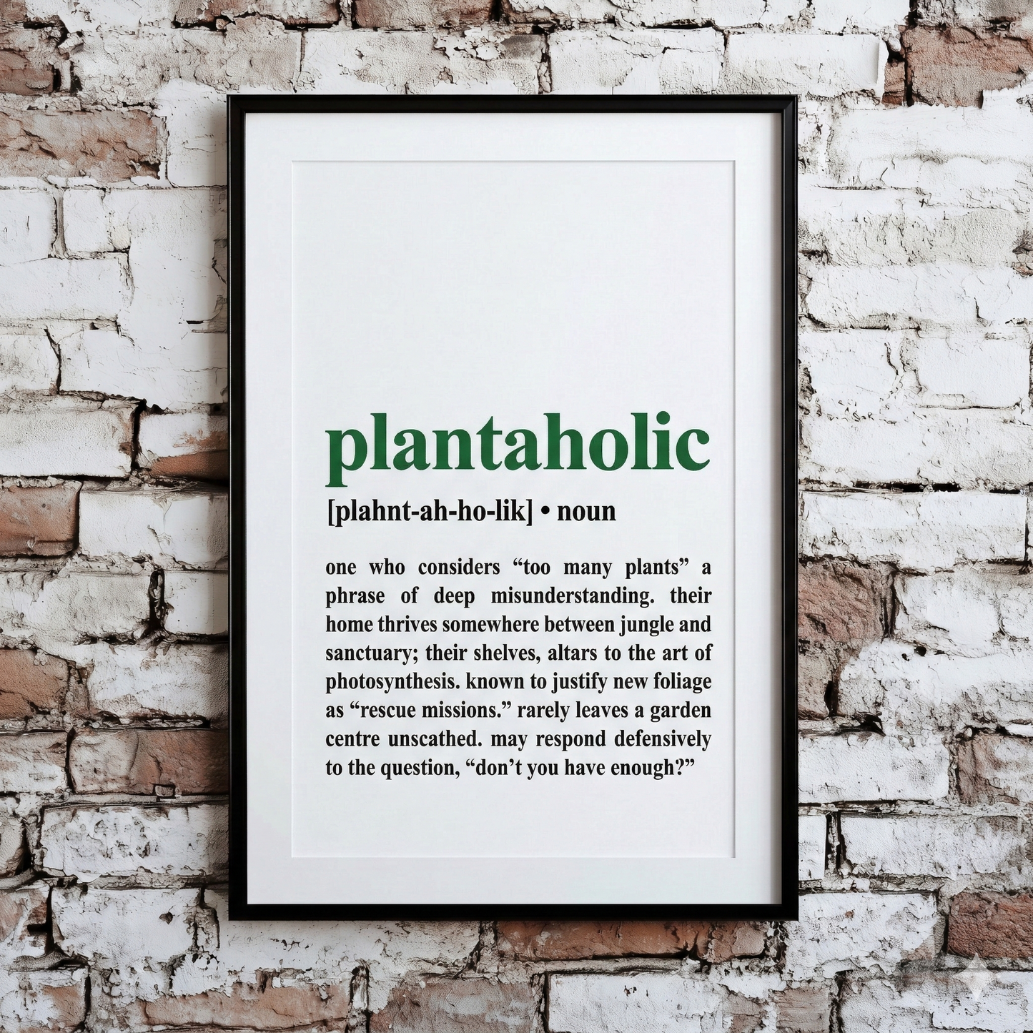 A Plantish print of the phrase "Plantaholic" in the style of a dictionary definition, showing the pronunciation against a white background, hanging on a brick wall.