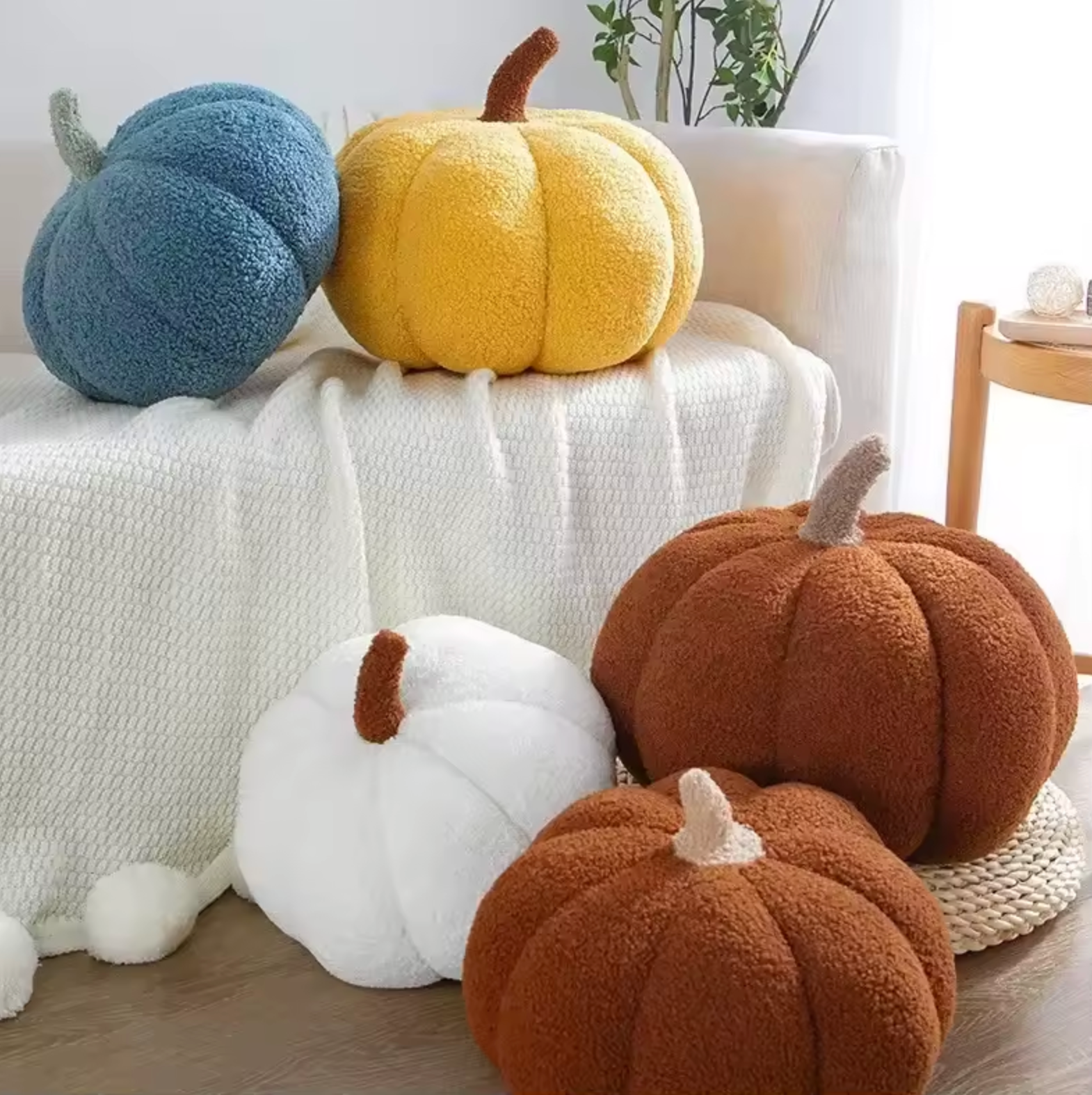 A collection of Plantish Pumpkin Plush Cushions shown in a bright living room. Some are on a white sofa and some are on a wood floor. 