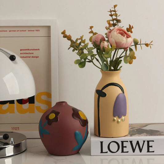 The Plantish Retro Floral Ceramic Vase shown in Sand and Mauve on a white table styled with a lamp. The Sand vase is on a white book and has peonies inside. 
