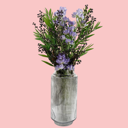 Side view of the Plantish Ridged Vase, made of clear glass, highlighting the vertical ridge design. Shown with artificial flowers inside and on a block pink background.