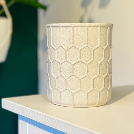 A small white ceramic plant pot with a raised honeycomb design and flecks of gold colour, on top of a white wood shelf. 