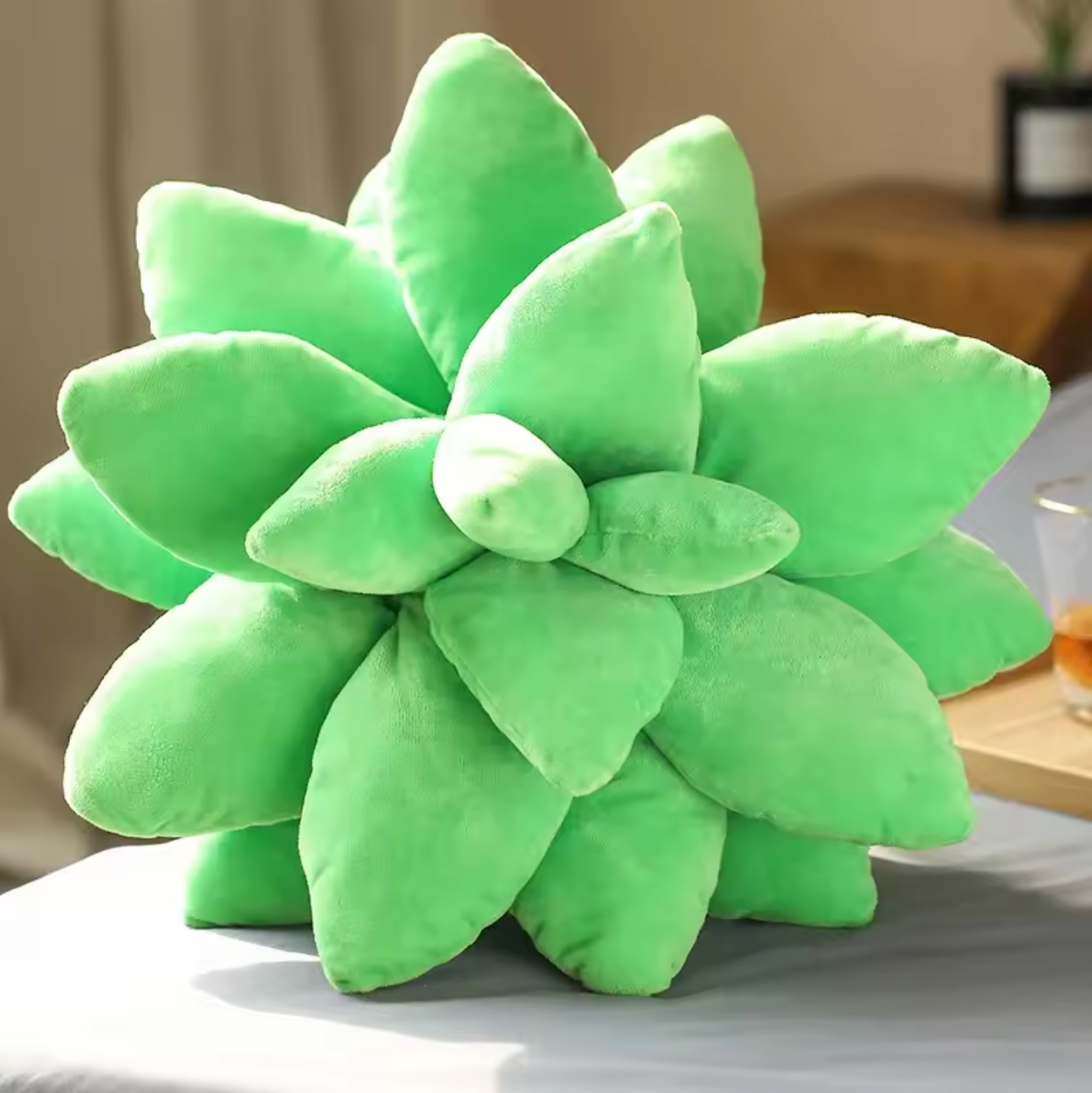 A close up of the top of the Plantish Succulent Plush Cushion in Bright Green, showing the soft leaves, placed on a grey bed with a bedroom in the background. 