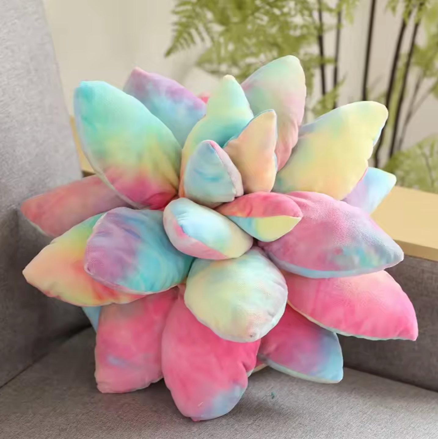 A close up of the top of the Plantish Succulent Plush Cushion in the Multicolour design, showing the soft leaves, placed on a grey sofa. 