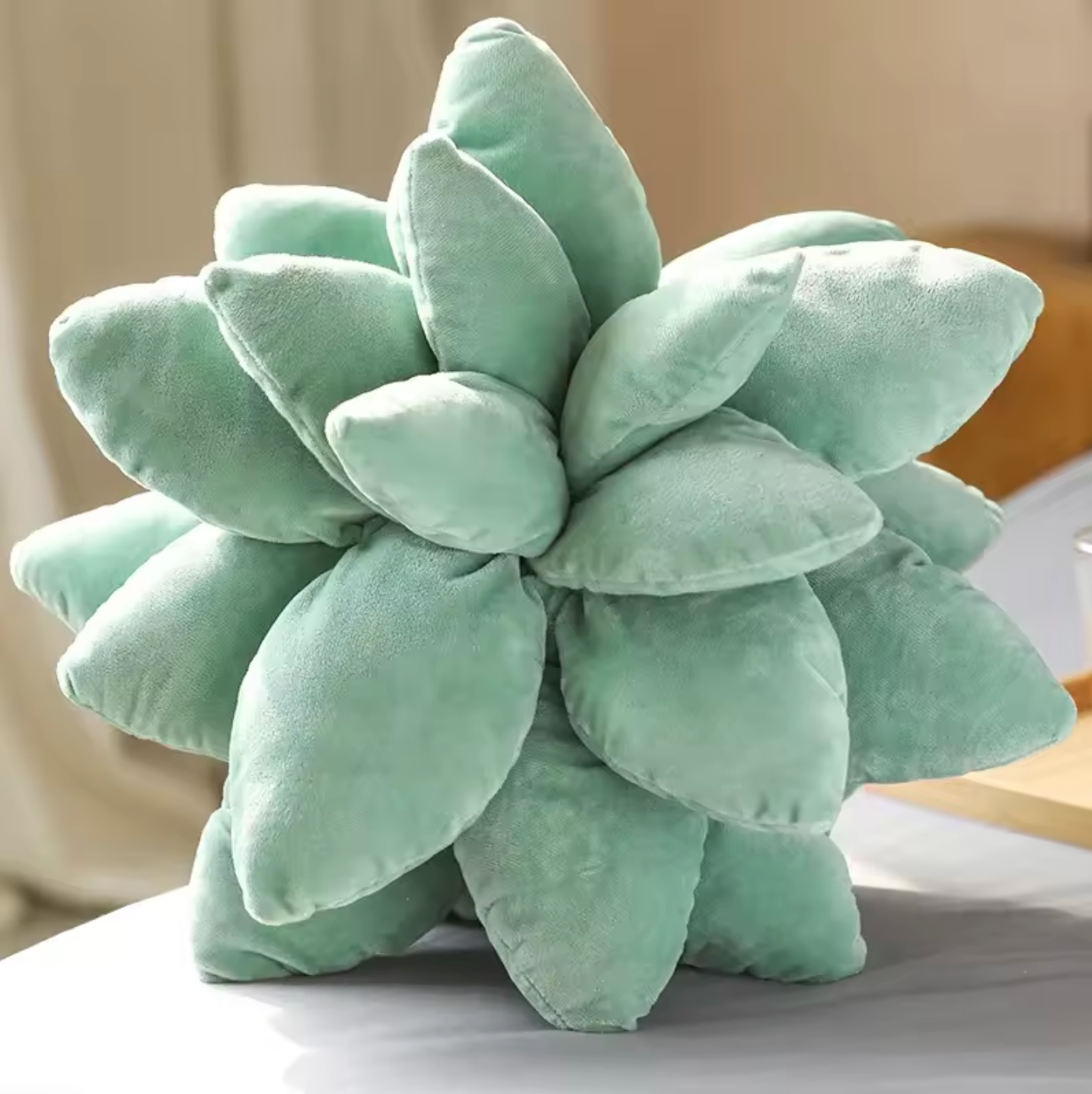 A close up of the top of the Plantish Succulent Plush Cushion in Sage Green, showing the soft leaves, placed on a grey bed with a bedroom in the background. 
