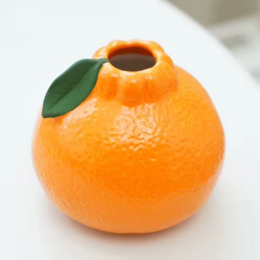 A close up of the Plantish Tiny Orange Vase on a white table. The dimpled orange  detailing and single painted green leaf are visible, as well as the opening of the vase.  