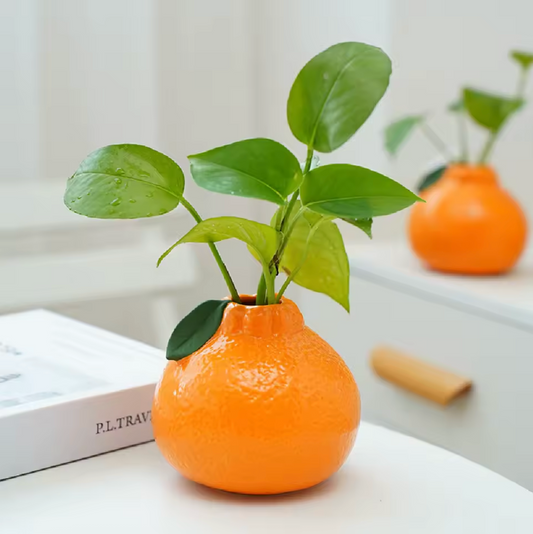 The Plantish Tiny Orange Vase shown in a bright corner of a house on a white table that also has a white book on it. There is a green ivy plant stem inside the vase. 