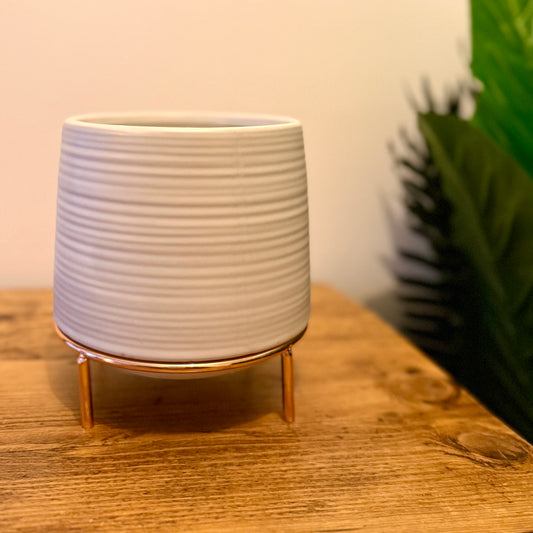 A white ceramic plant pot with horizontal grooves and tapering sides which is on top of a rose gold coloured metal stand, placed on a wood table. 
