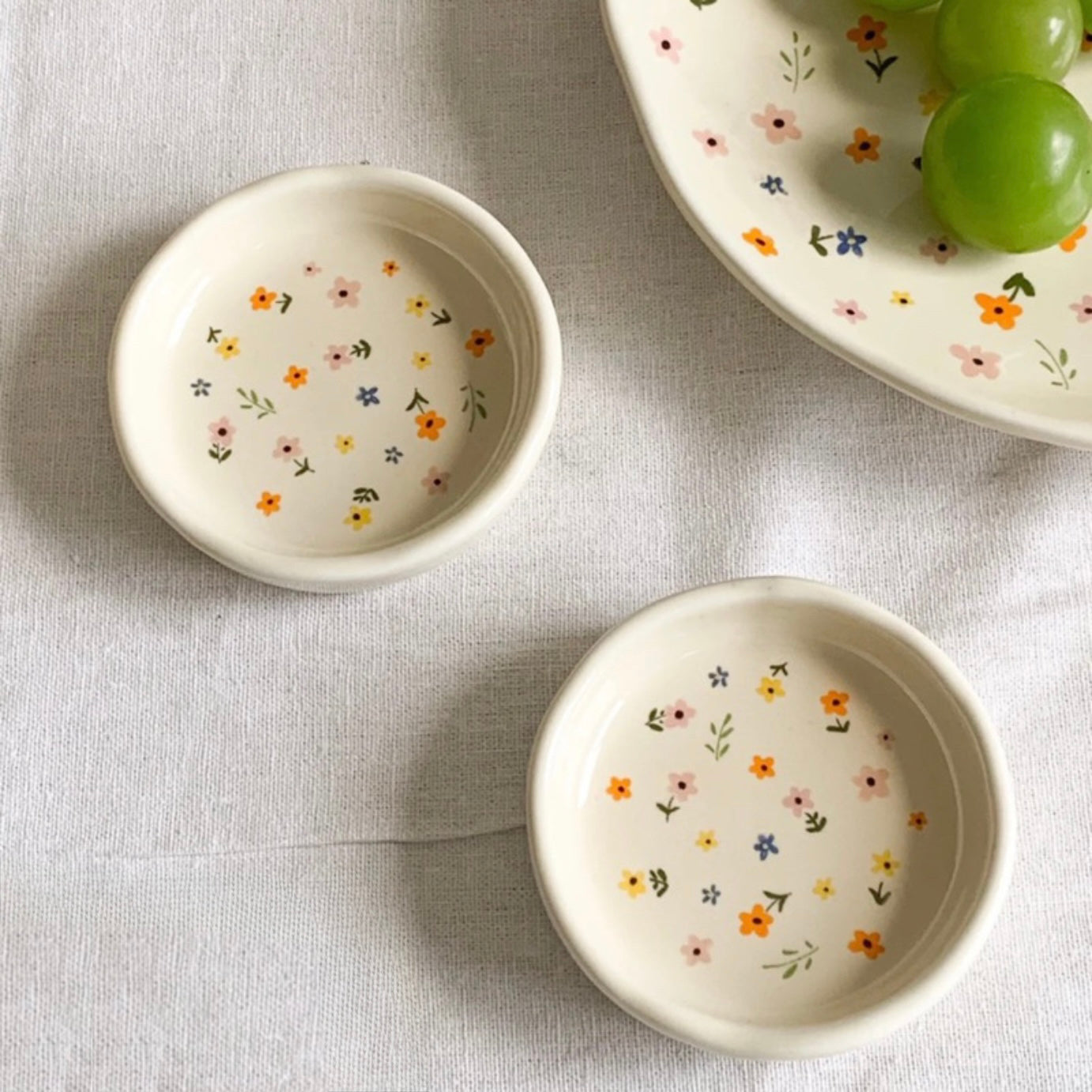 A close up of two Plantish Wildflower Sauce Pots on a linen tablecloth, along with a wildflower Plate in the corner of the image.