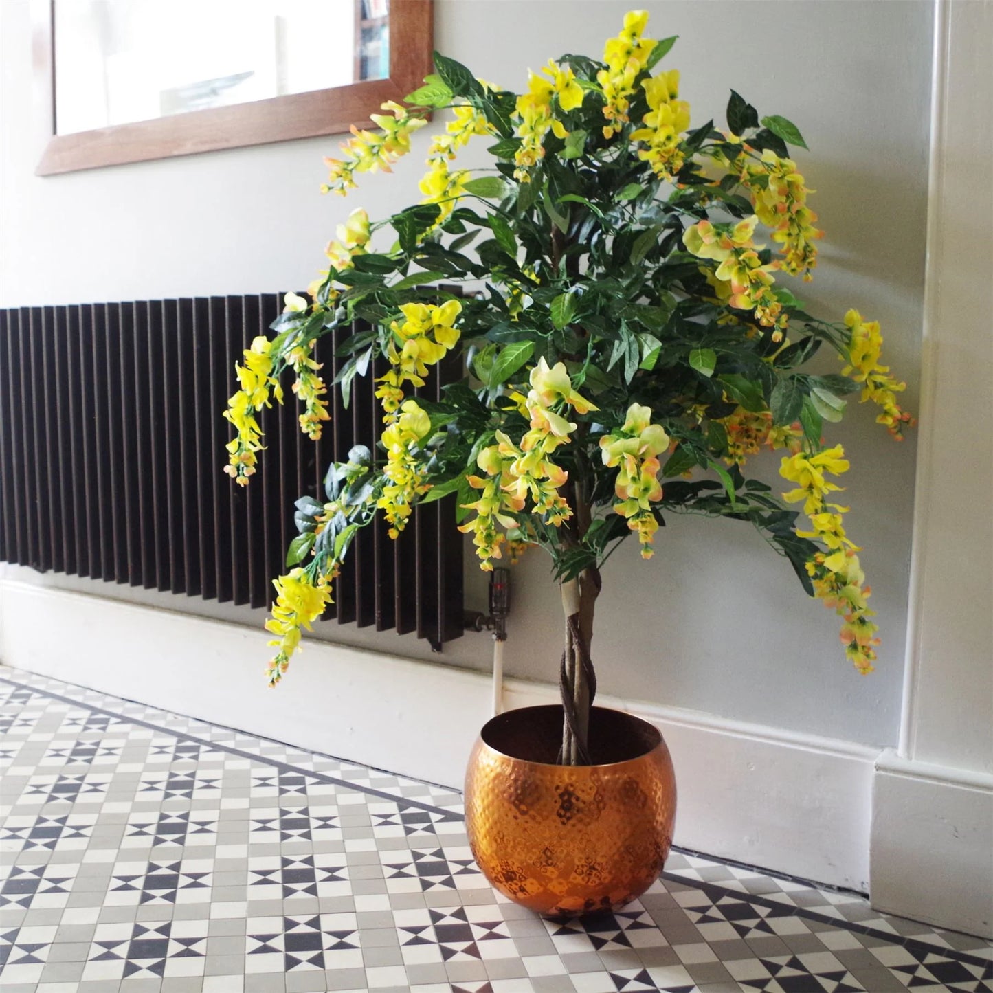 The Plantish artificial wisteria with yellow flowers and green leaves shown inside a copper hammered pot placed inside a bright hallway with a mosaic floor. 