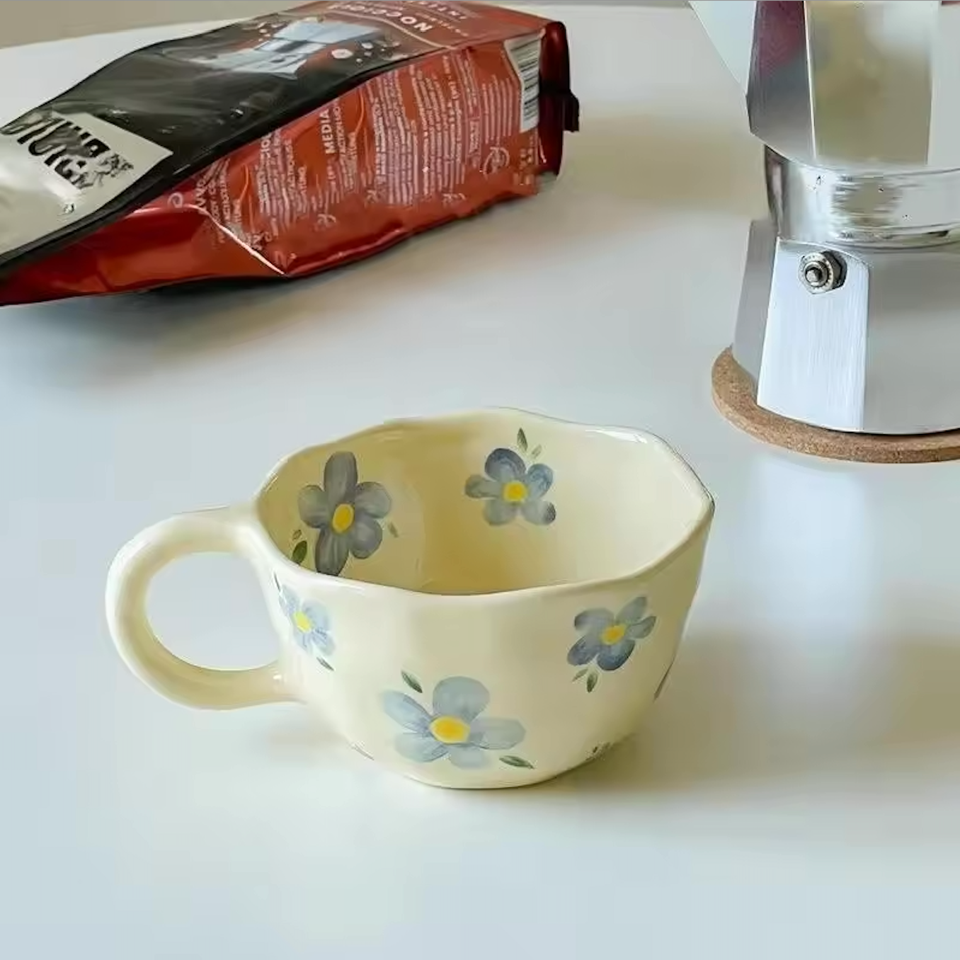 A close up image of the Blue Flower design of the Plantish Wonky Flower ceramic mug, on a white table that also has a stovetop coffee pot and a bag of coffee. 