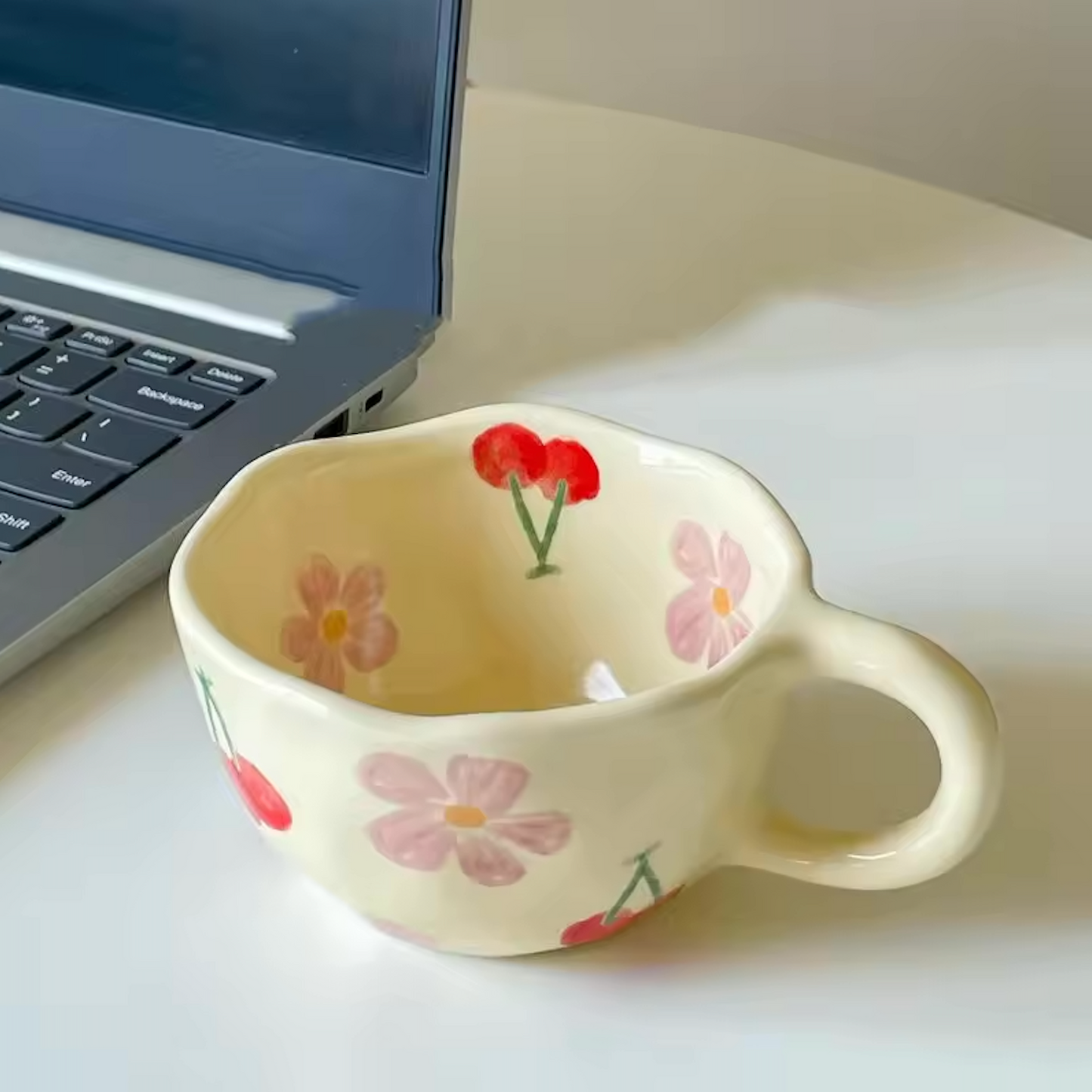 A close up image of the Cherry Blossom design of the Plantish Wonky Flower ceramic mug, placed on a white table that also has a laptop on it.