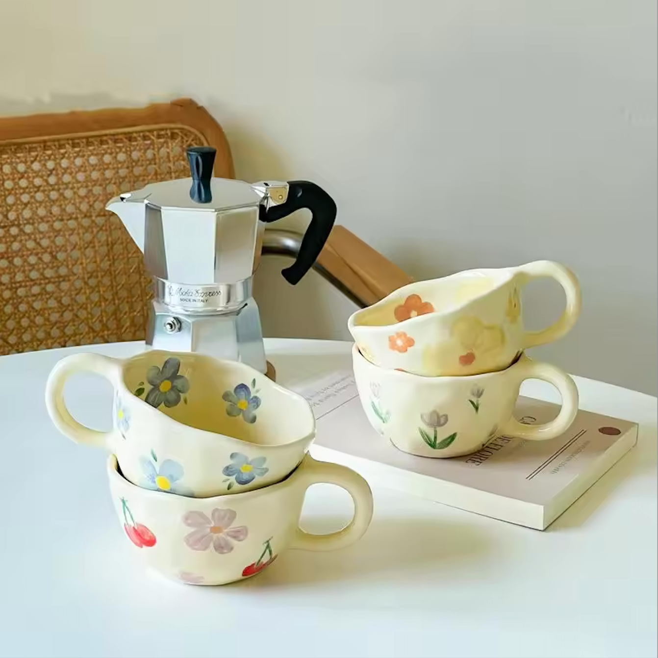 An image of four Plantish Wonky Flower Mugs on a white table with a stovetop coffee pot and a book. There is a rattan chair behind between the table and wall.