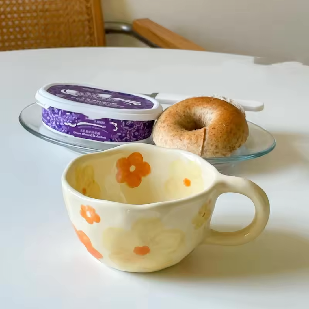 A close up of the Orange Flower design of the Plantish Wonky Flower ceramic mug on a white table with a bagel and pot of cream cheese, and a rattan chair behind.