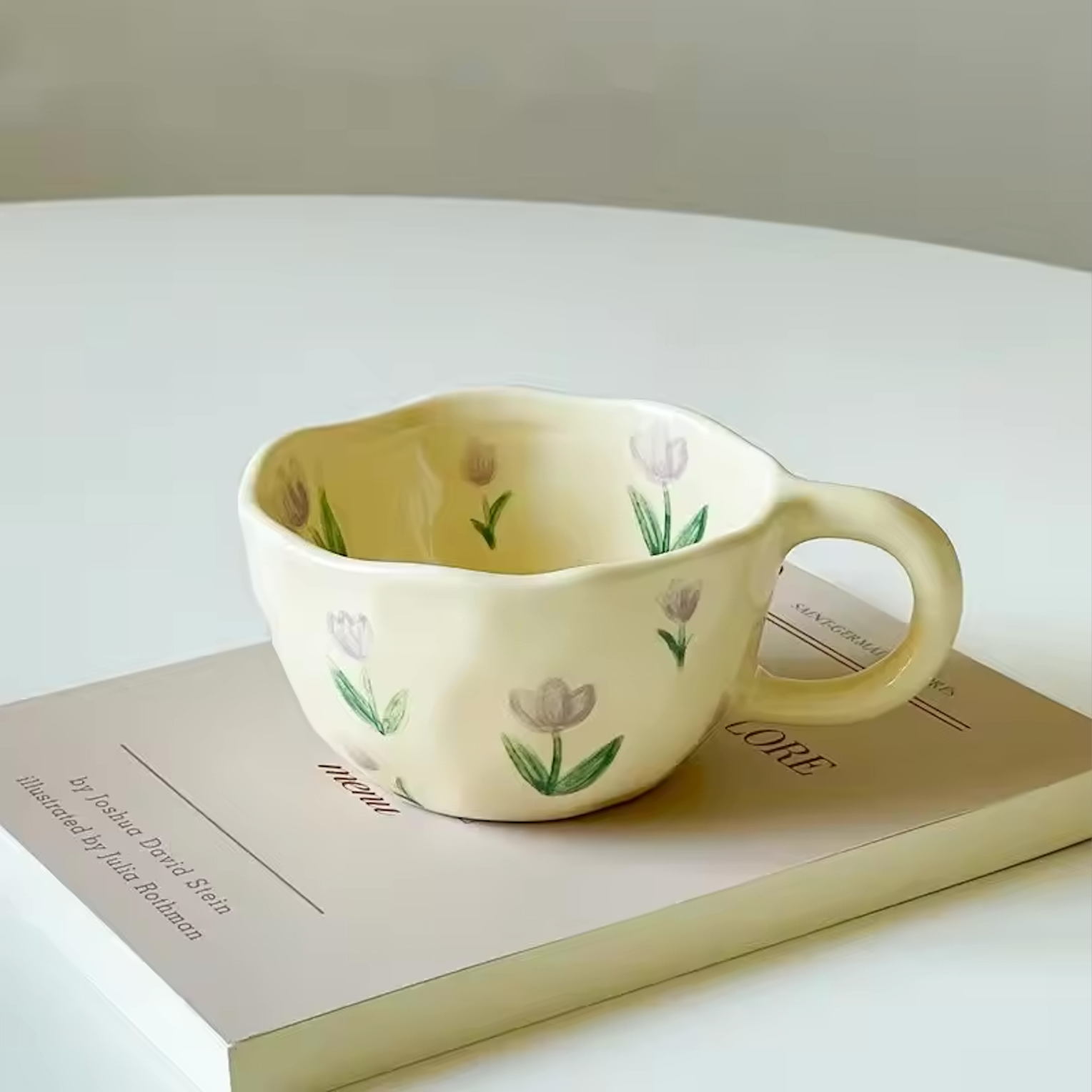 A close up image of the Purple Tulip design of the Plantish Wonky Flower ceramic mug, placed on a book which is resting on a white table. 