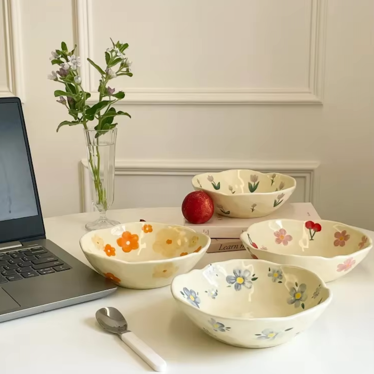The Plantish Wonky Flower Plate shown in all four variants on a white table, along with other items: a laptop, books, a clear vase. There is a white panelled wall in the background. 