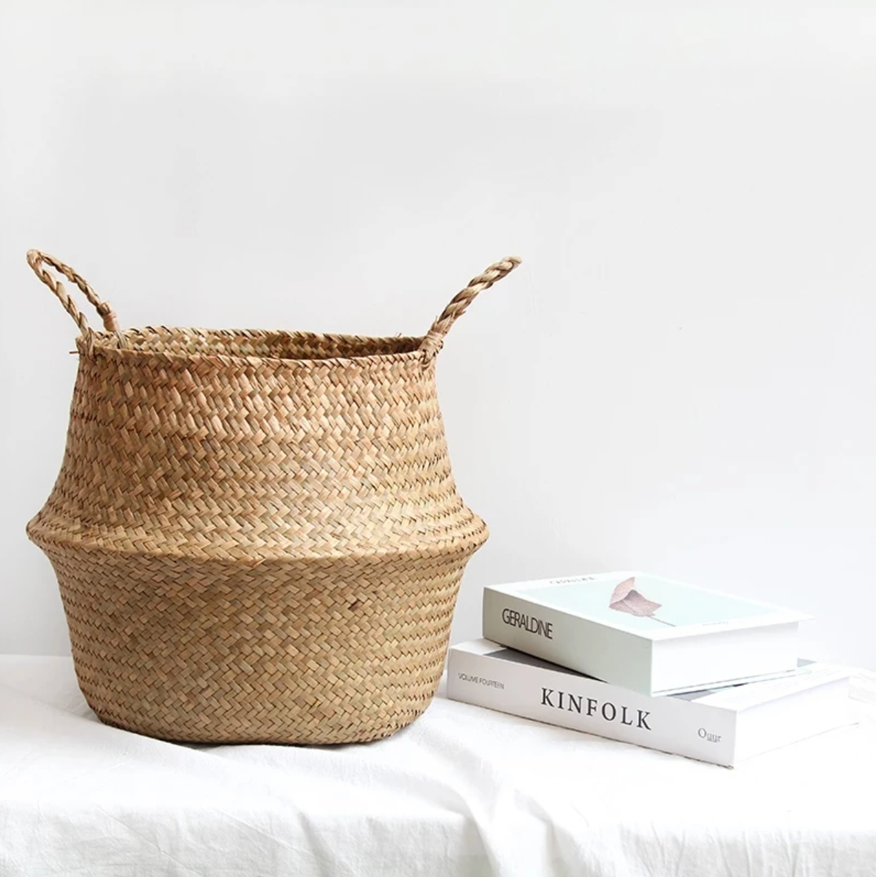 The Plantish Woven Wicker Basket fully extended, and positioned on a white bed alongside two hardback books. There is a white wall in the background.