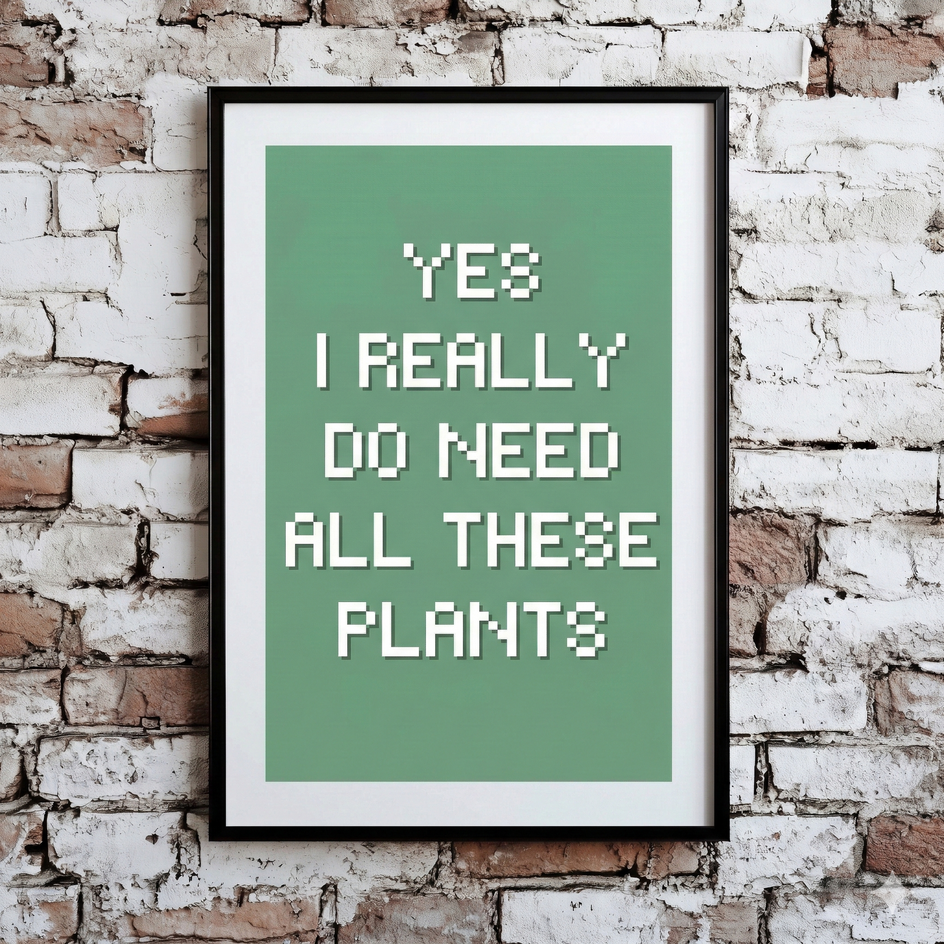 A Plantish print showing the phrase "Yes I Really Do Need All These Plants" in a white font against a green background, hanging on a brick wall.
